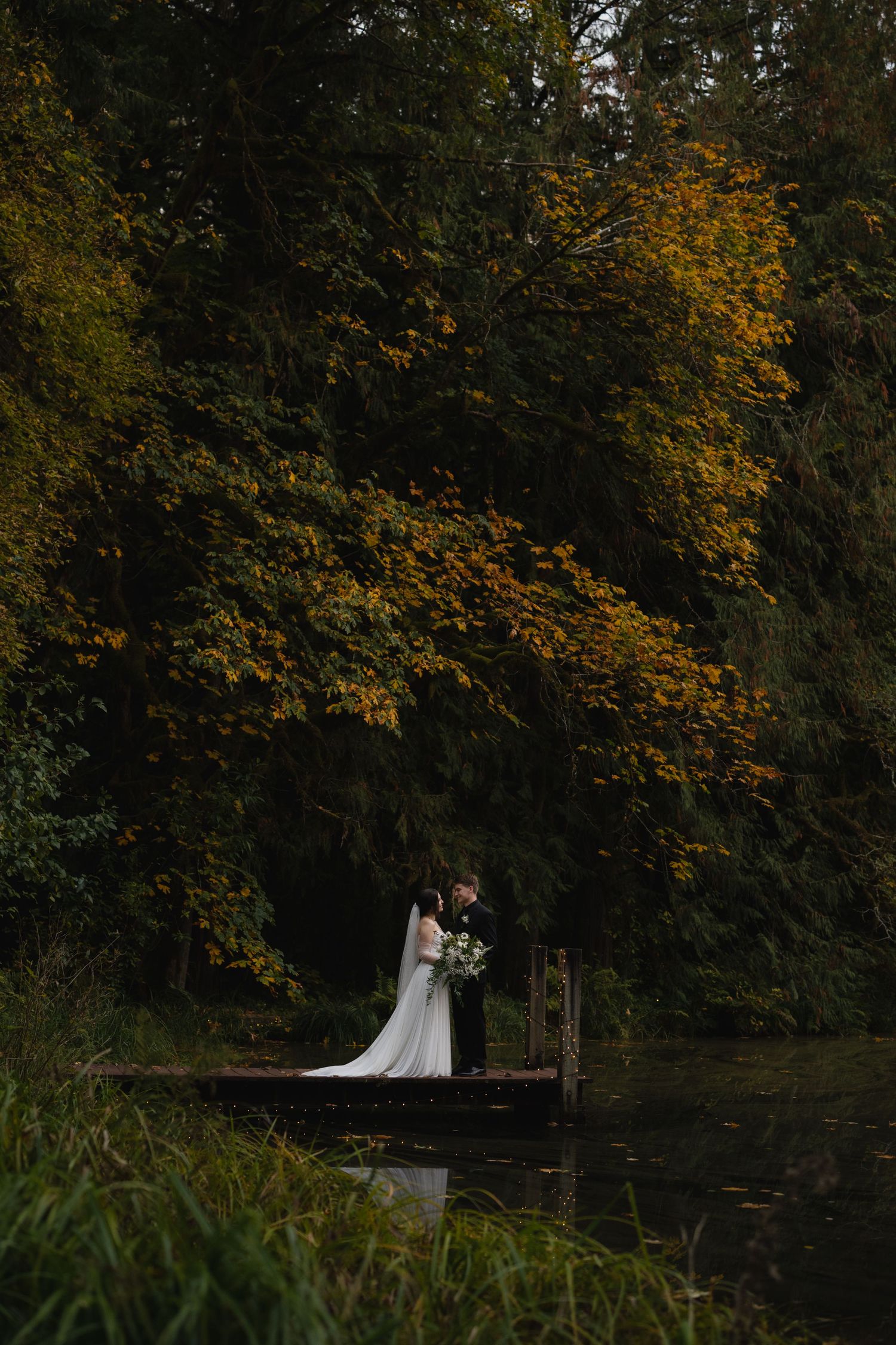 A couple shares an intimate moment on a wooden dock surrounded by autumn foliage and water.