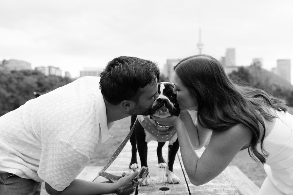 A sweet couple smiles at each other with a puppy between them in a heartwarming black and white outdoor photo.