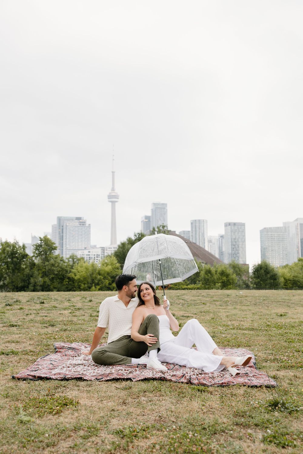 A couple shares a romantic moment under a clear umbrella on a blanket with the Toronto skyline in the background.
