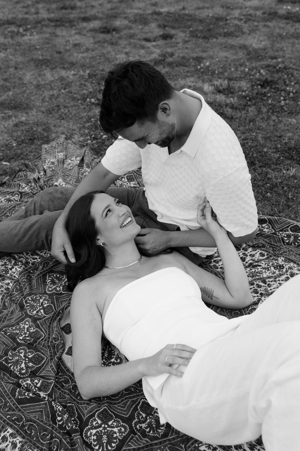 A romantic couple lies together on a patterned blanket outdoors in a series of intimate black and white portraits.