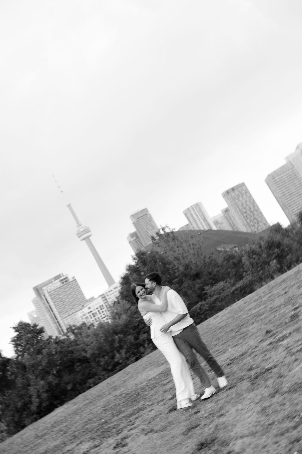 A couple embraces on a grassy hill with the Toronto skyline and CN Tower visible in the background on an overcast day.