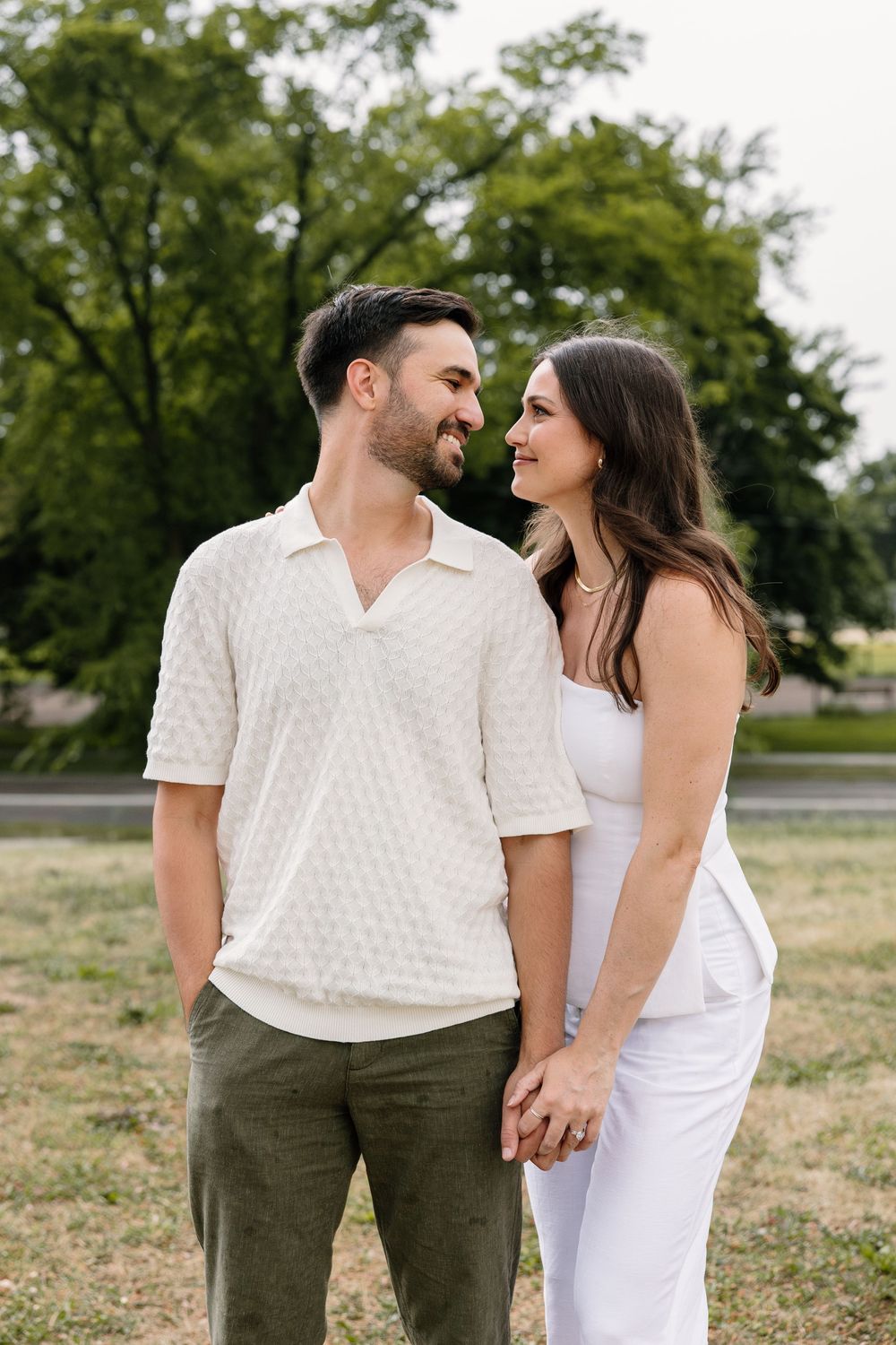 A couple in white and olive outfits share joyful moments in a grassy outdoor setting during a casual photoshoot.