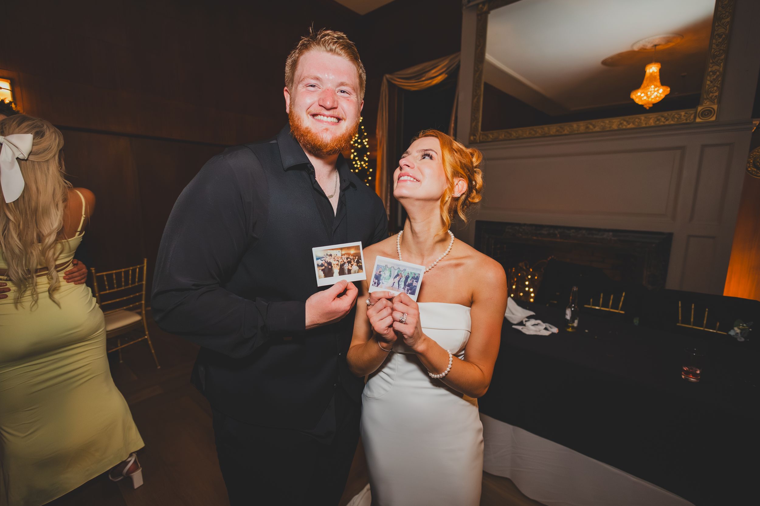 Bride and groom in  formal attire smile and hold up polaroids at an indoor celebration event.