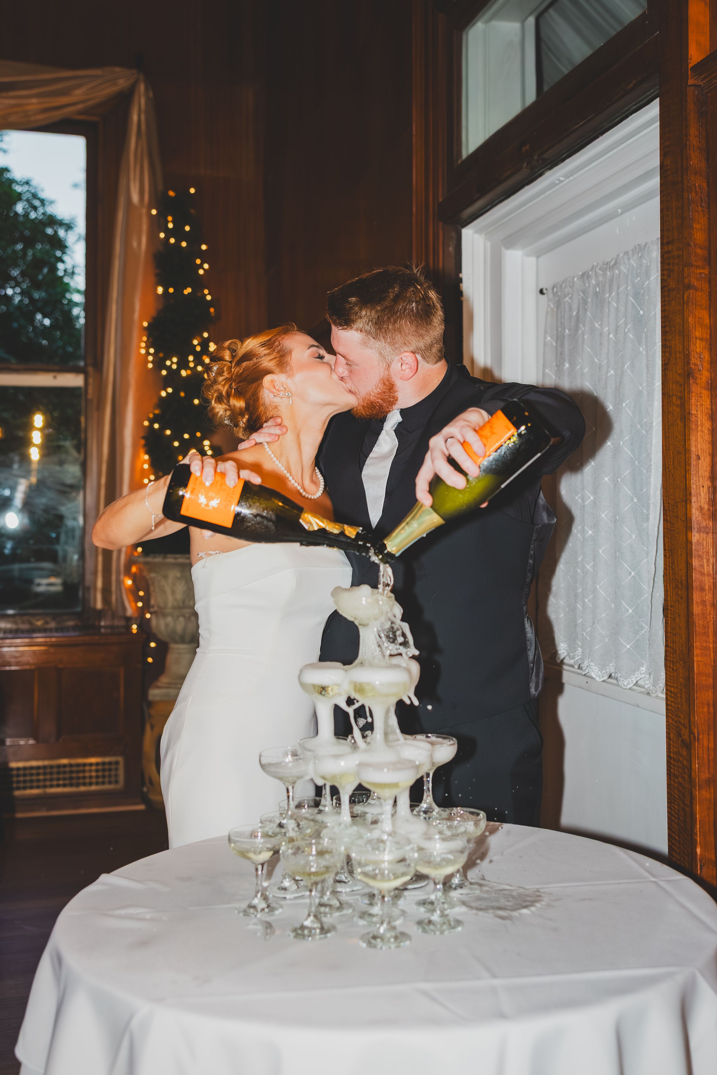 Bride and groom perform a romantic champagne tower pour at their wedding reception, with glasses stacked on white tablecloth.