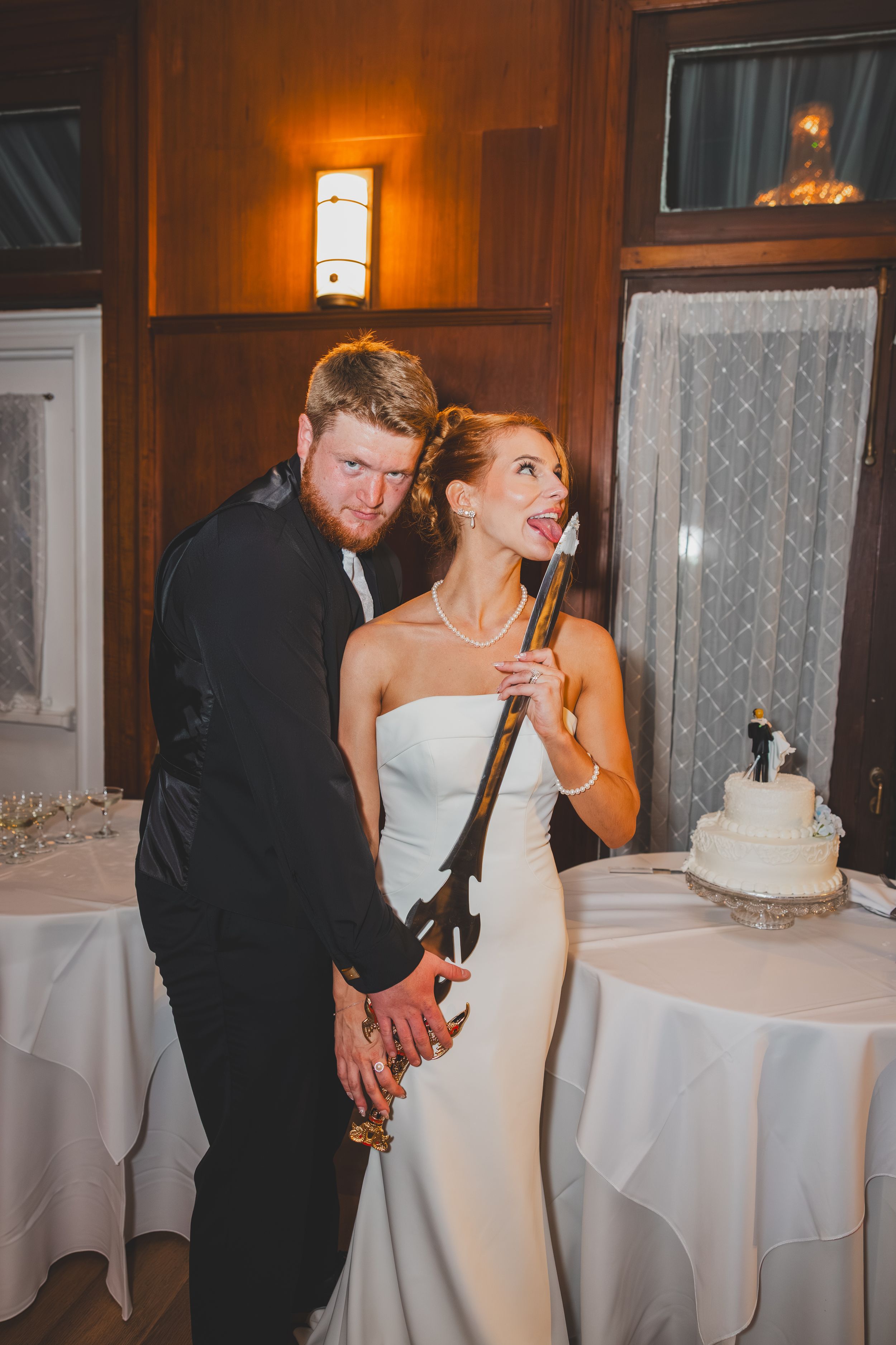 A bride in a white gown cuts her wedding cake with a sword while other guests mill around tables in a warmly lit reception hall.