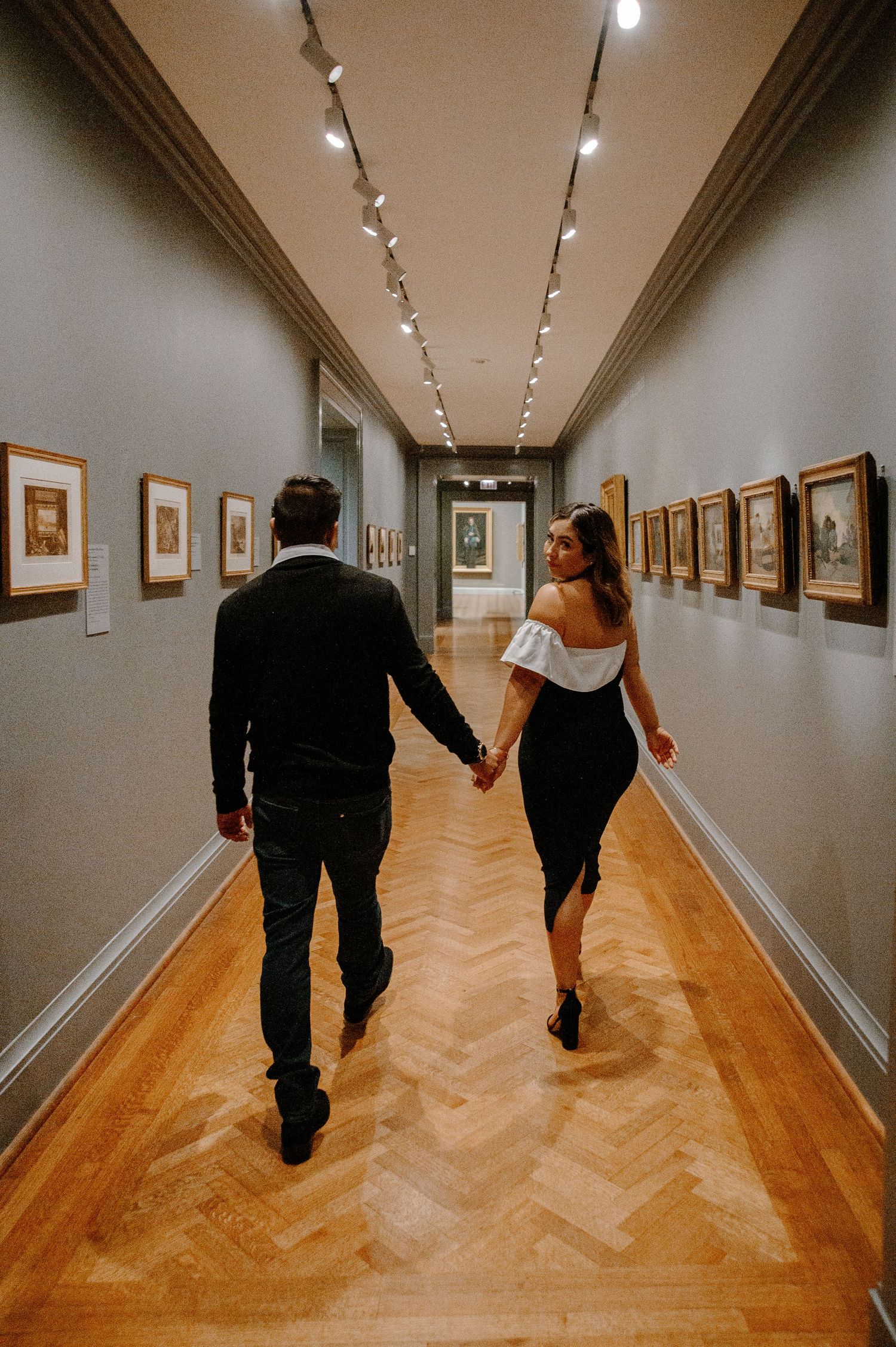 Two people walking through an art gallery hallway with wooden floors and framed artwork lining the white walls.