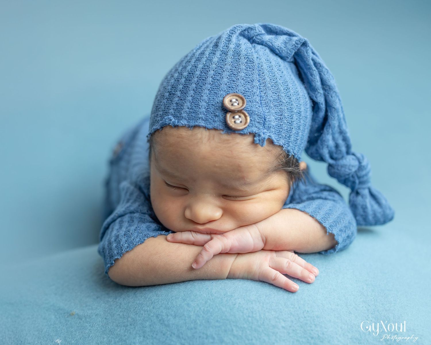 Sleeping newborn in soft blue knit hat rests peacefully on cozy blanket with tiny hand under cheek.