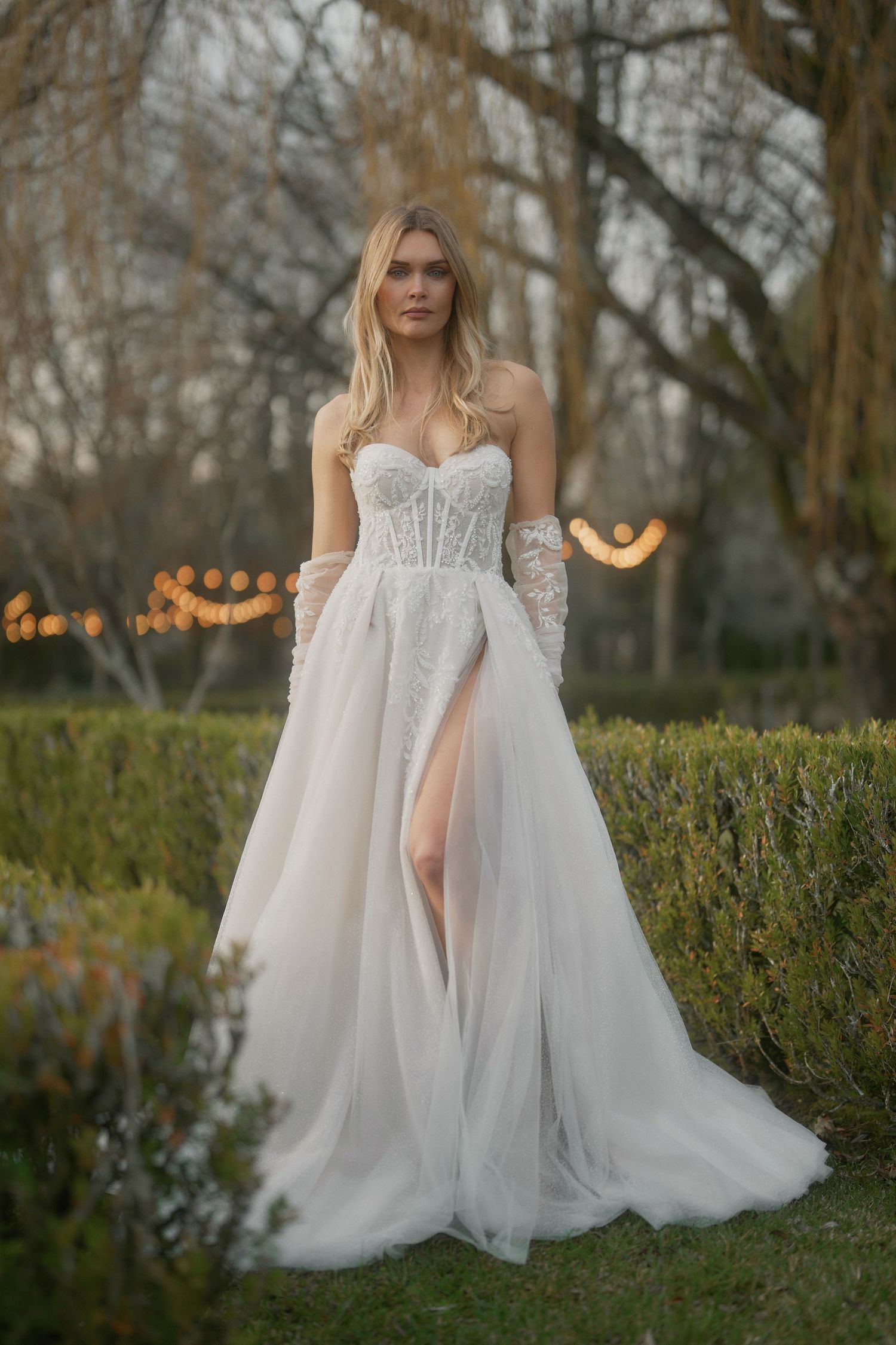 A romantic garden wedding dress with lace bodice, tulle skirt and high slit stands elegantly among trees at dusk.