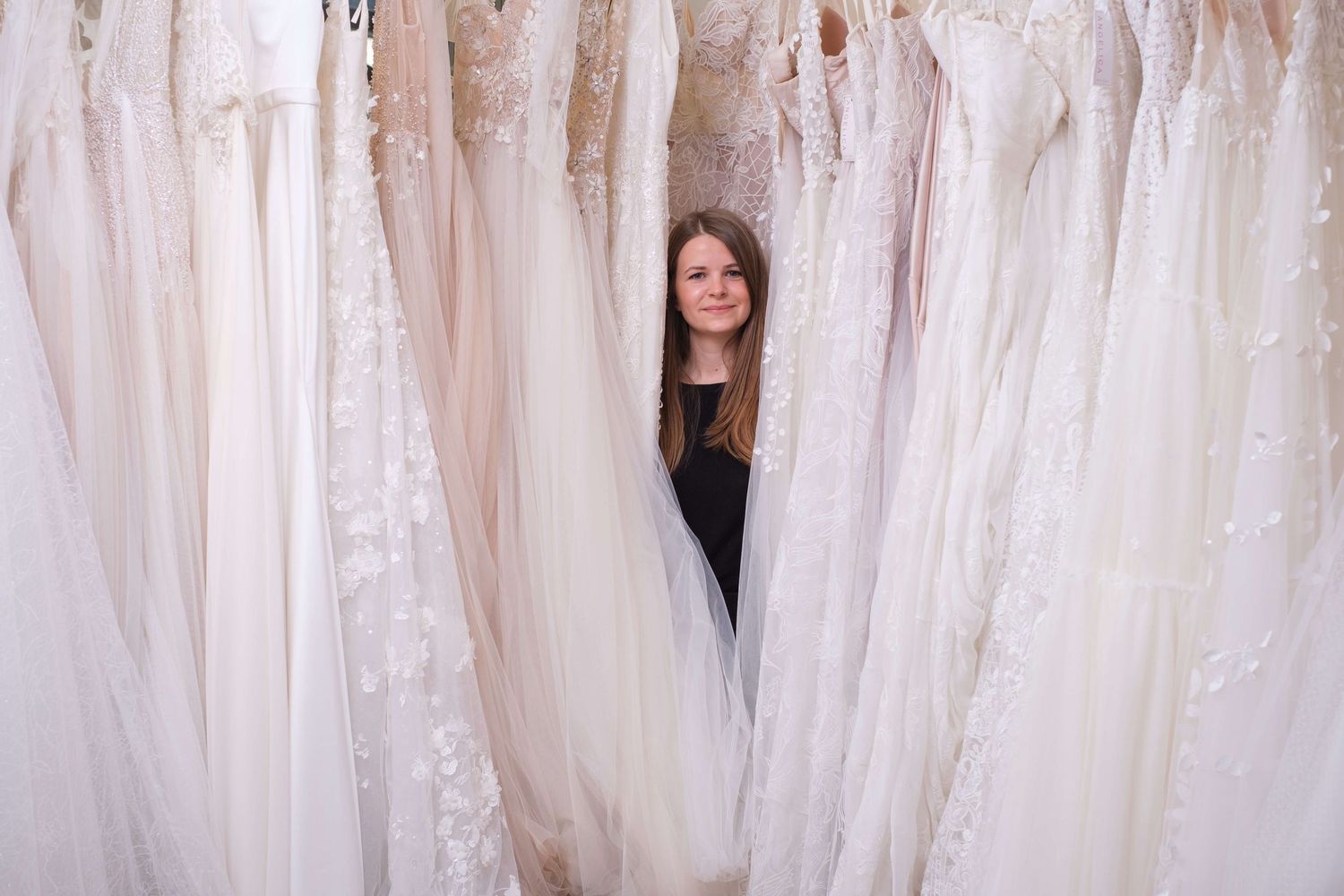 Between rows of white wedding dresses hanging in a bridal shop, someone peeks through while wearing a black top.
