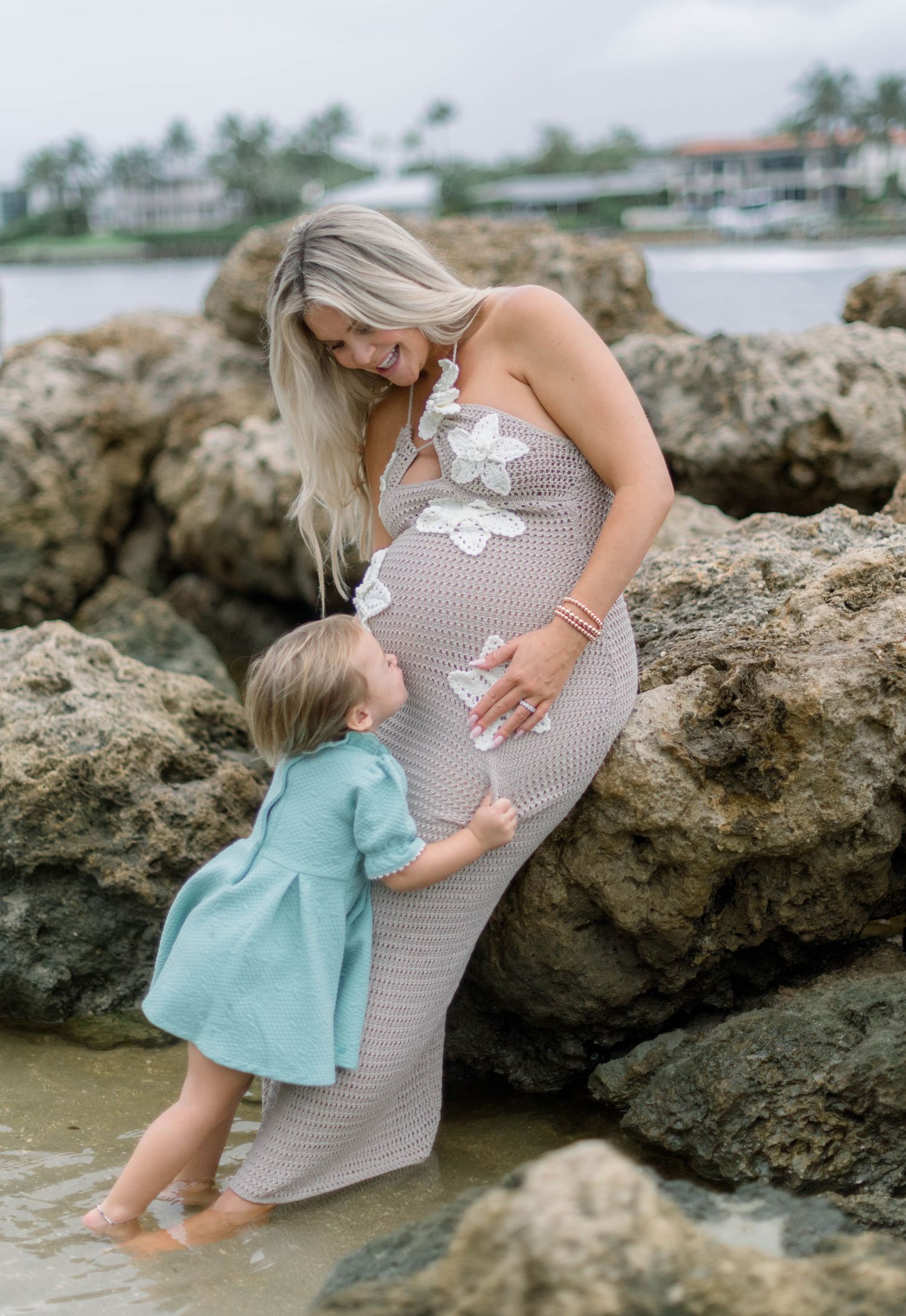 Mother and child share tender moment on rocky beach with ocean and palm trees in background.
