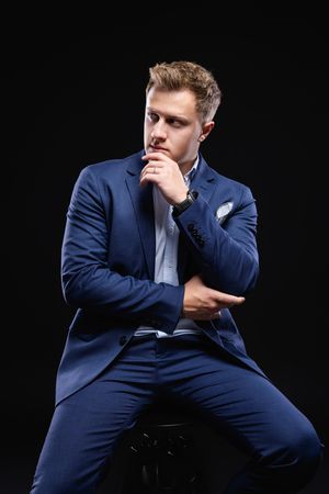 Professional portrait of man in blue suit posing thoughtfully on black background.