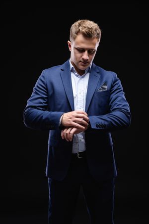 Man in navy suit adjusts sleeve cuff against black background.