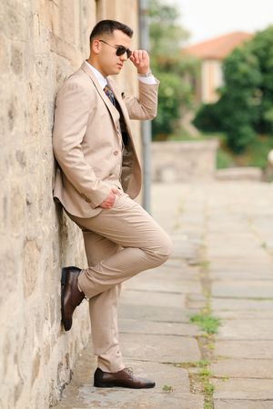 A stylish person in a beige suit strikes various poses against a stone wall outdoors.