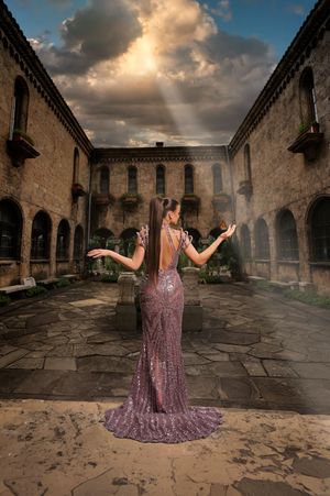 Woman in sequined gown stands in historic stone courtyard with dramatic sunbeams breaking through stormy clouds.