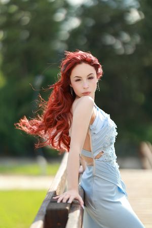 Woman with vibrant red hair in blue dress poses artistically outdoors.
