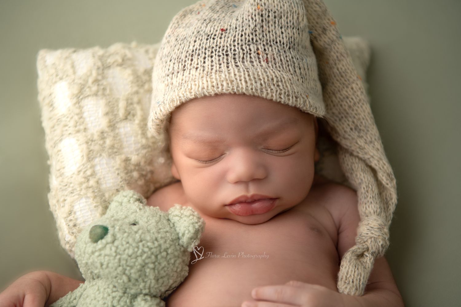Sleeping newborn wearing cream knit hat snuggles with mint green teddy bear on soft textured backdrop.
