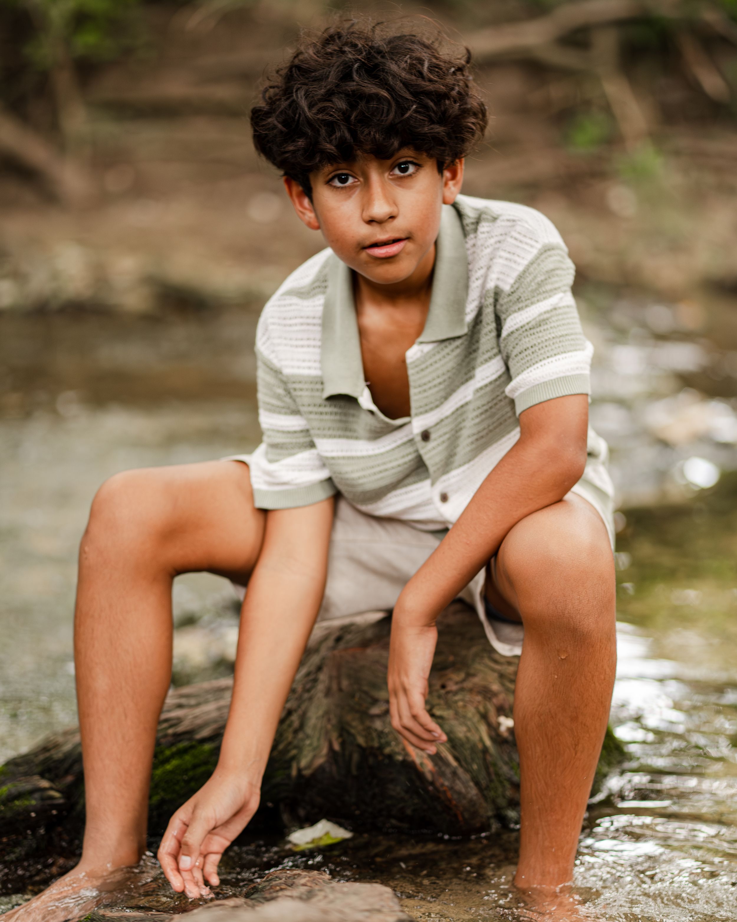 Person in a striped shirt sits on a rock by a stream, dipping fingers in the water during a tranquil outdoor moment.