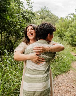 Two people sharing a joyful embrace while standing on a dirt path surrounded by lush greenery on a cloudy day.