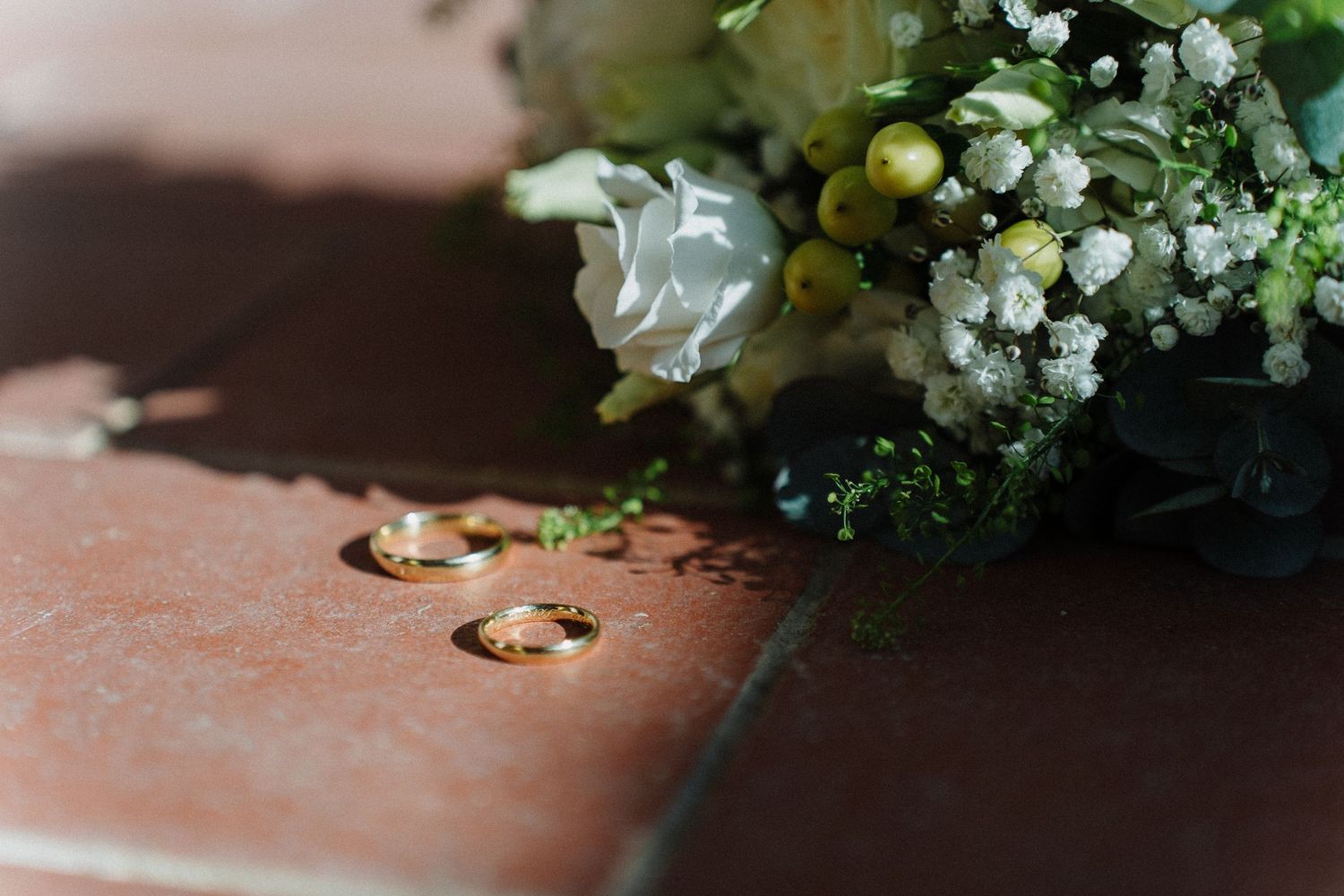 Two gold wedding rings resting on a wooden surface beside a white and green floral bouquet arrangement.