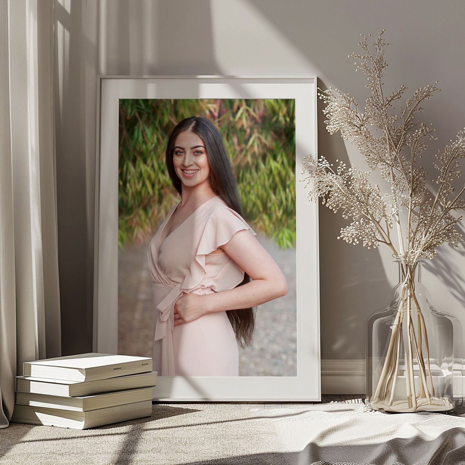 Framed portrait photo displayed on white table with stacked books and dried floral arrangement in sunlit room.