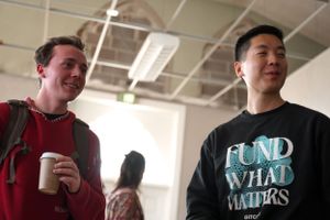 Two people having a conversation in front of a white wall, one wearing a red backpack.