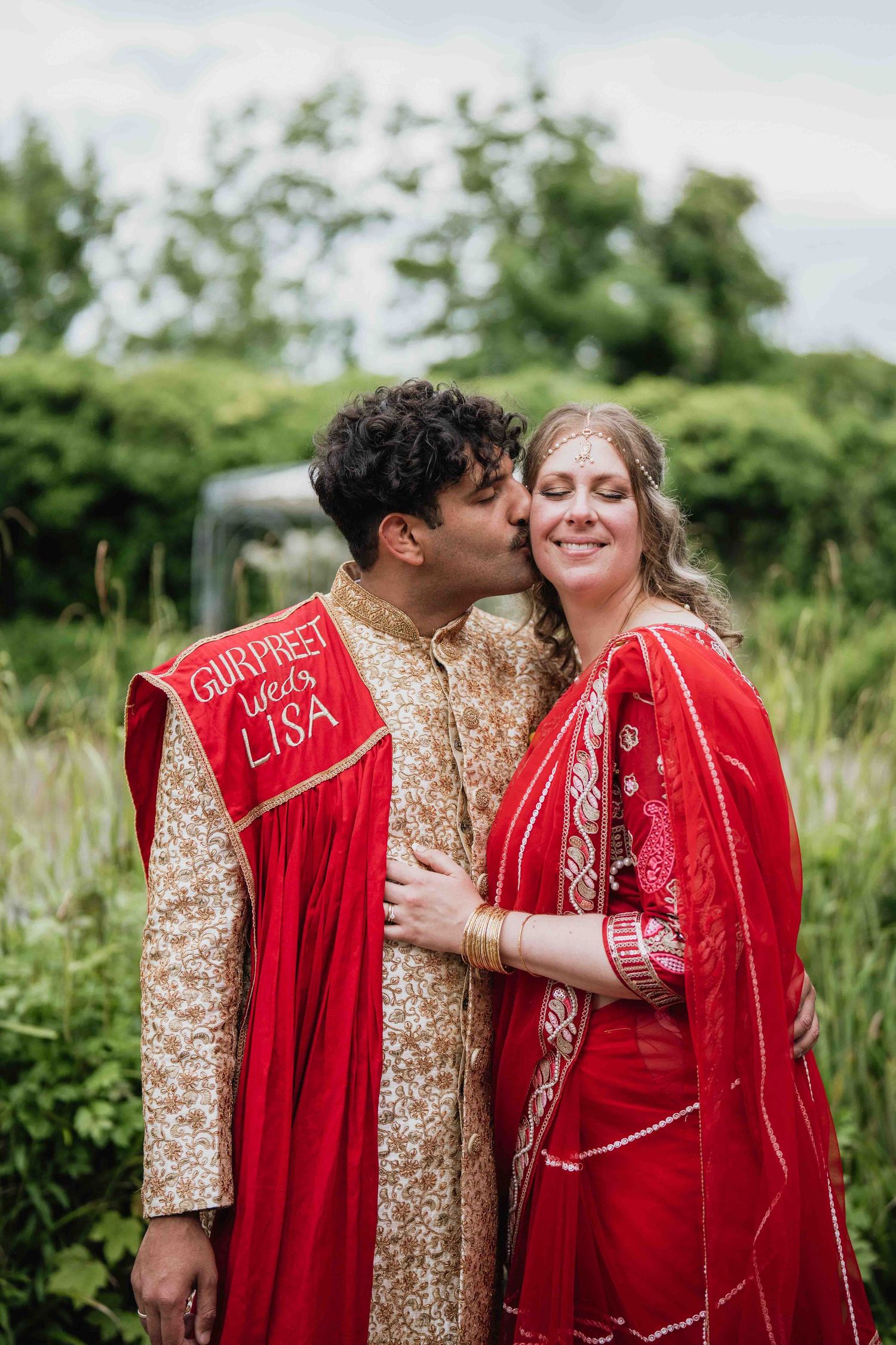 Wedding couple in traditional red and gold attire embracing in a lush garden setting.