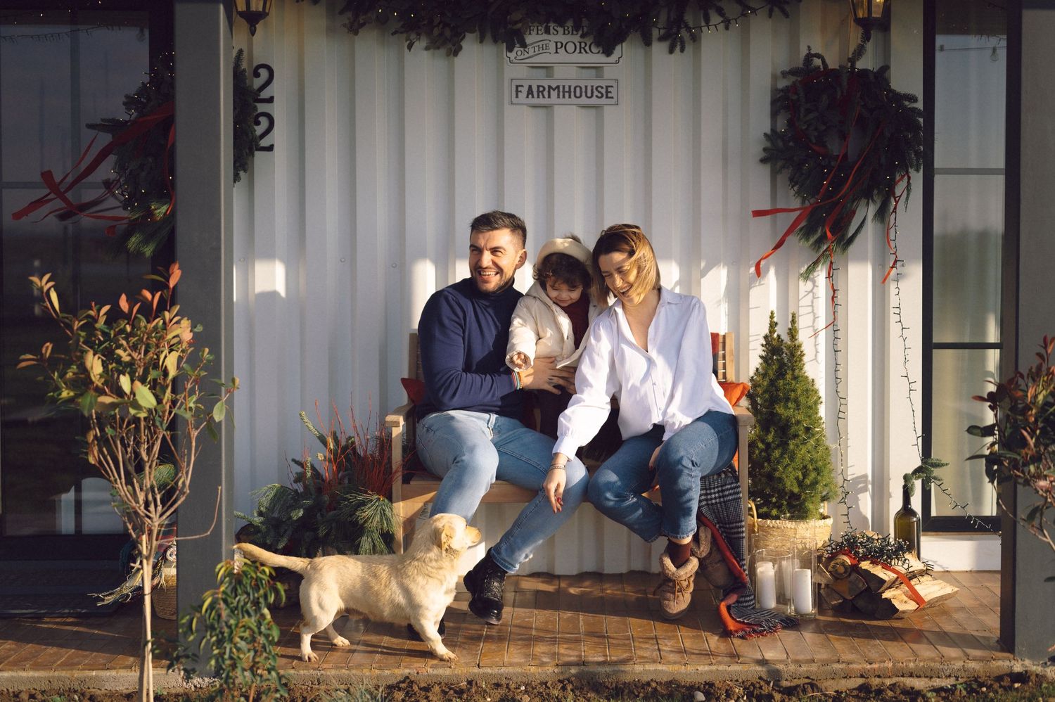 Two people relaxing on a cozy porch swing with their cat in front of a white house decorated with holiday greenery.