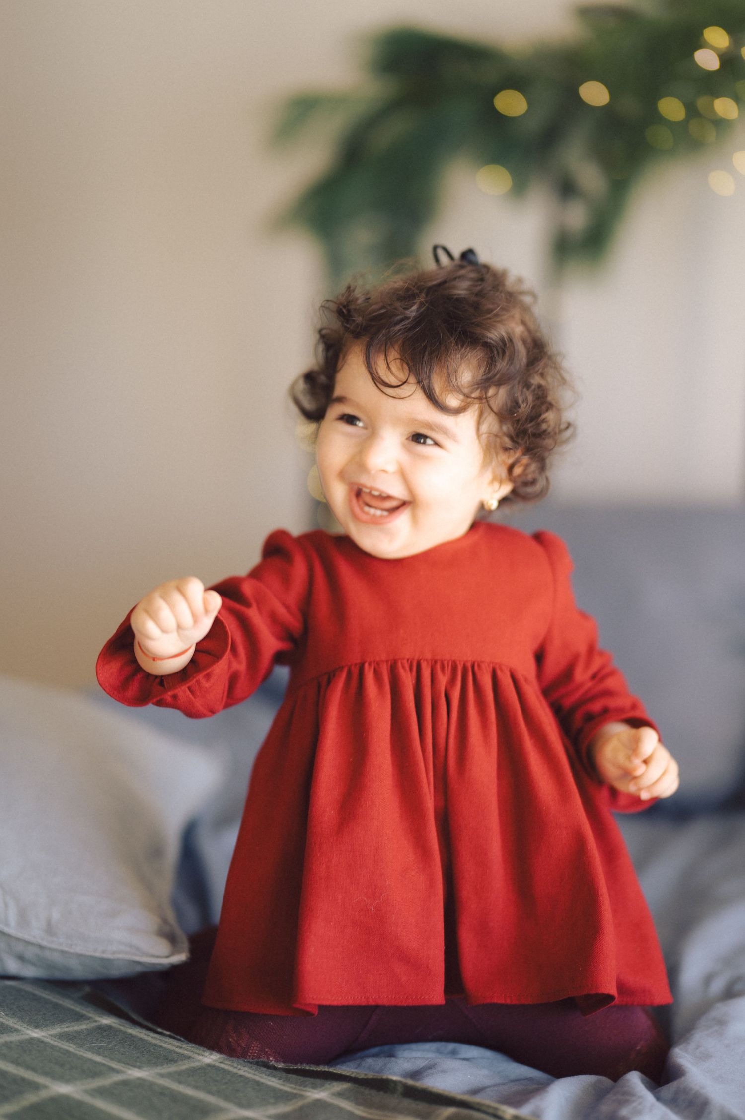 Toddler wearing red dress smiles while standing on gray bedding with holiday lights in background.