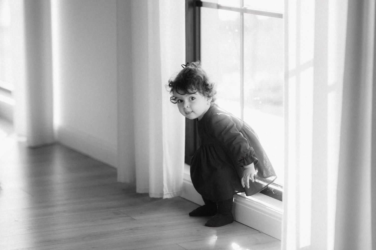Black and white photograph of a person in a dark sweater sitting by a window in a minimal white interior.