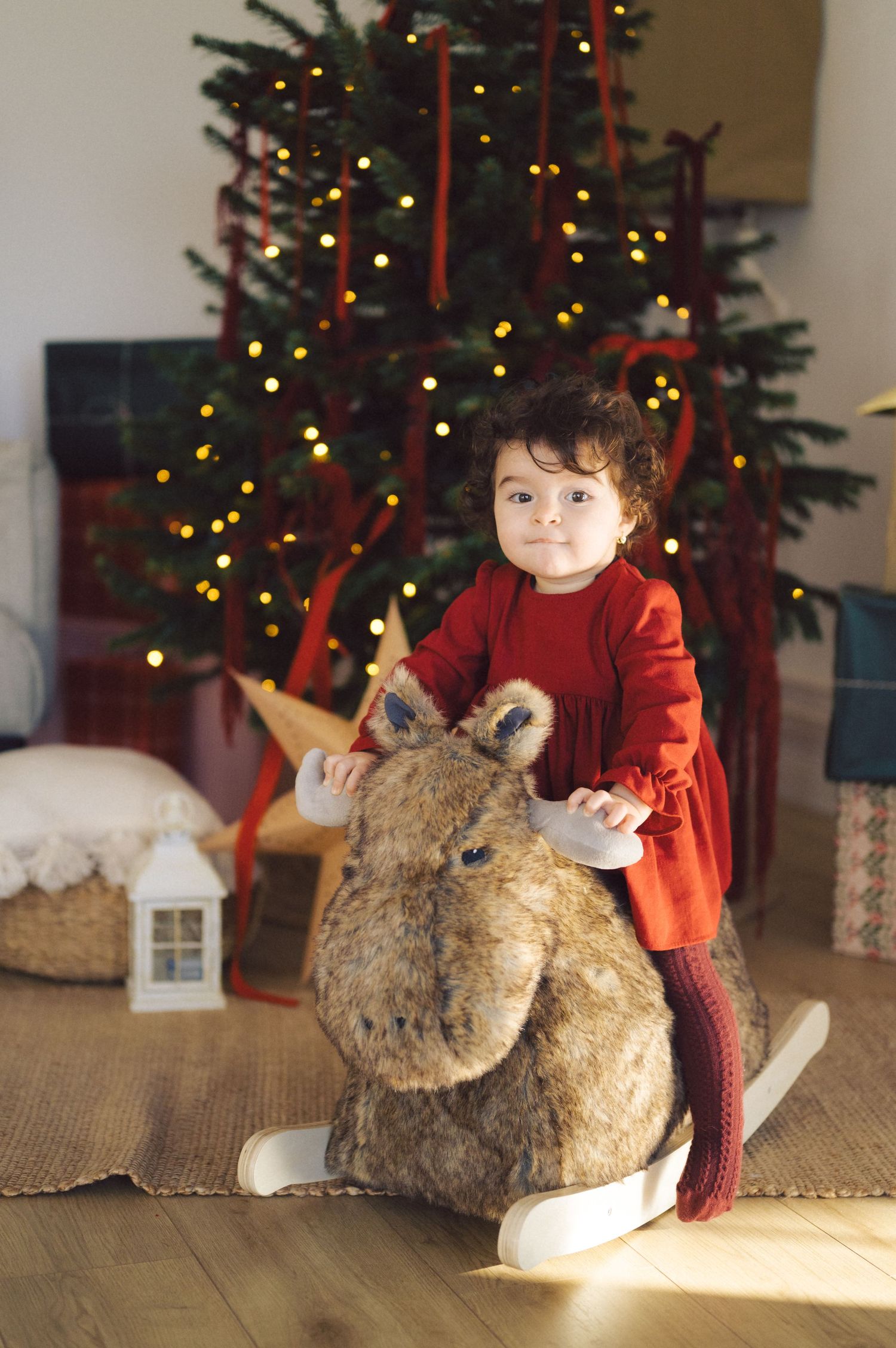 A child in red sits on the floor playing with a large plush reindeer next to a decorated Christmas tree.
