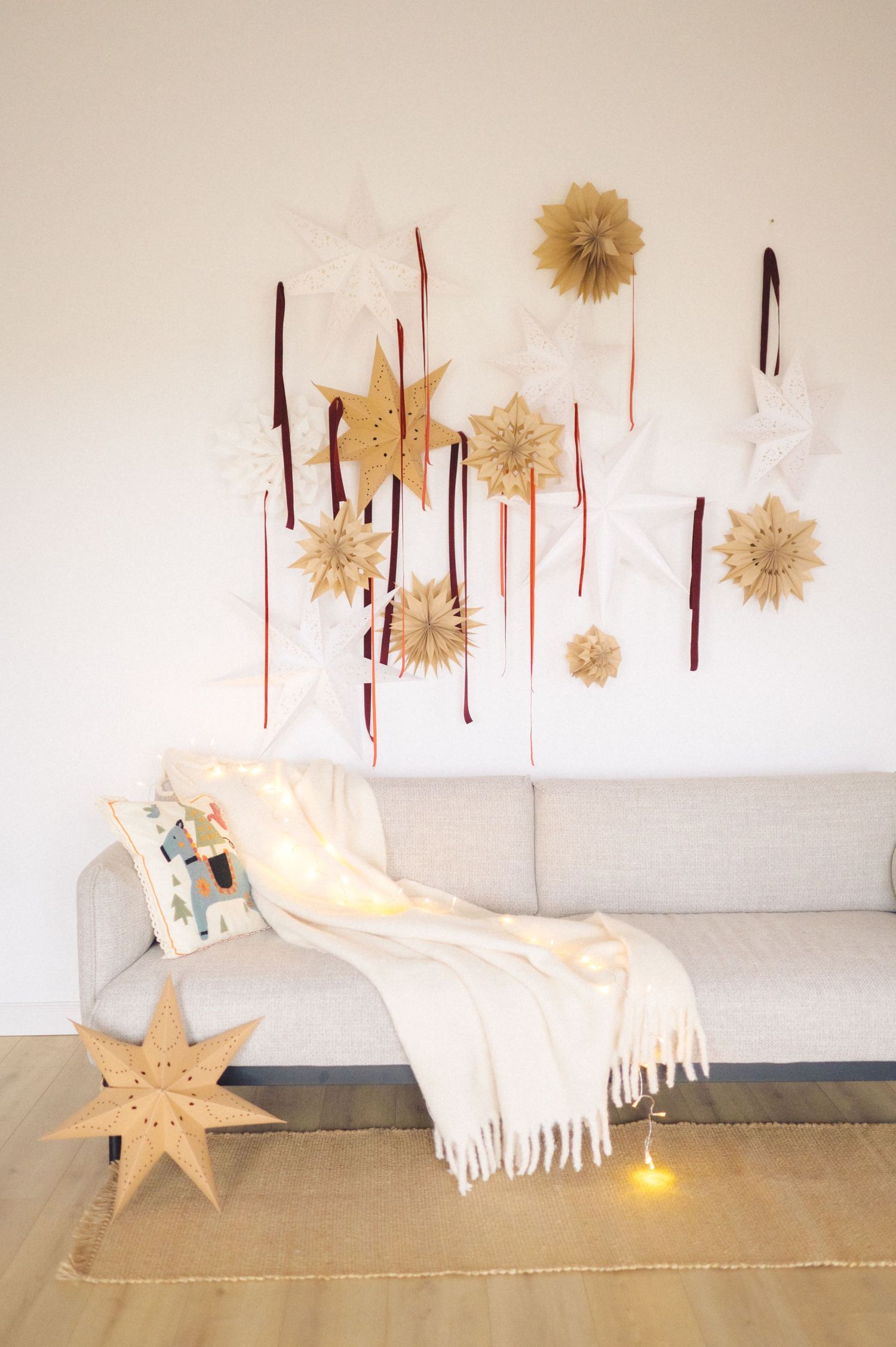 Minimalist living room with paper pompom wall decor, white sofa, cozy blanket, and illuminated star decoration on floor.