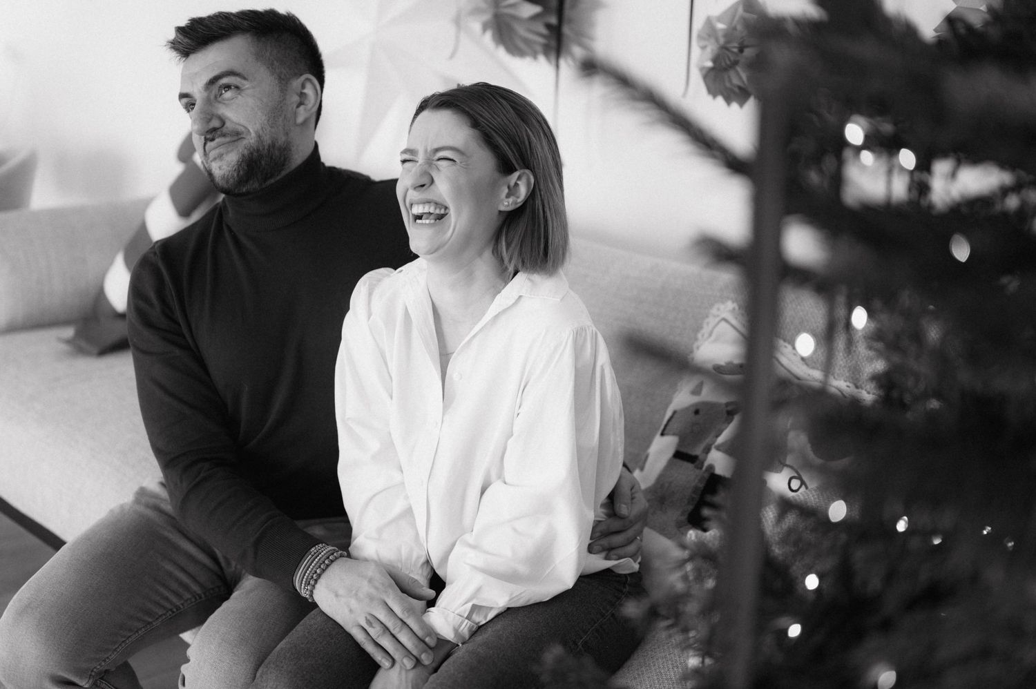 A couple shares a joyful moment together in black and white, sitting near twinkling holiday lights.