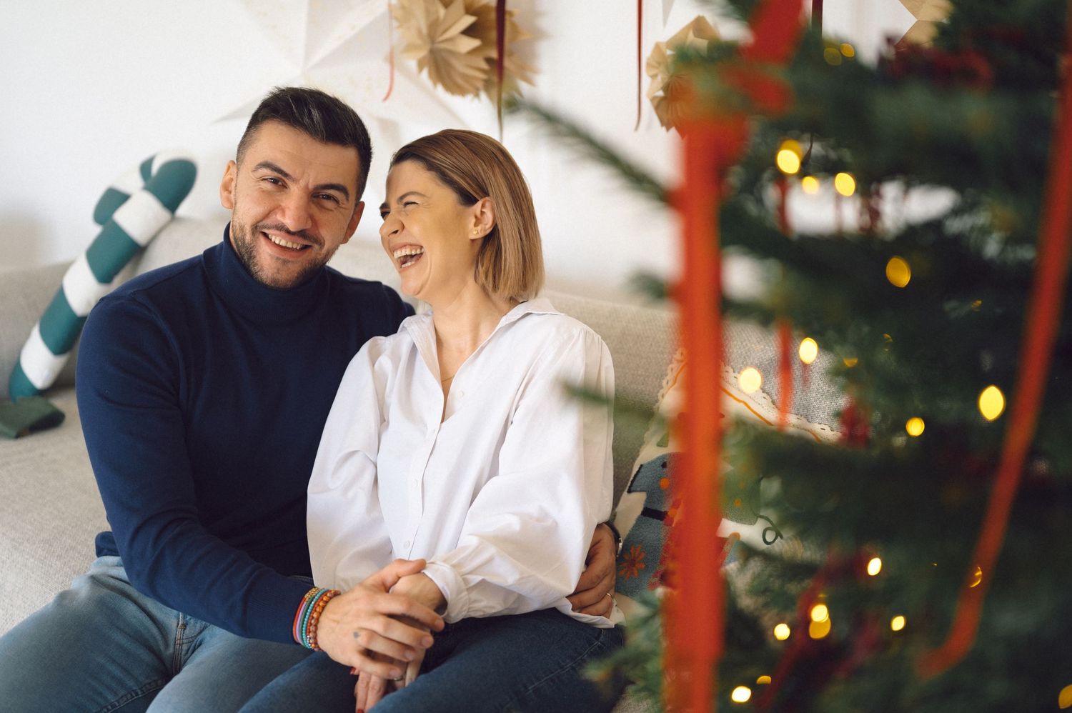 A joyful couple shares a laugh while sitting together by their decorated Christmas tree with twinkling lights.