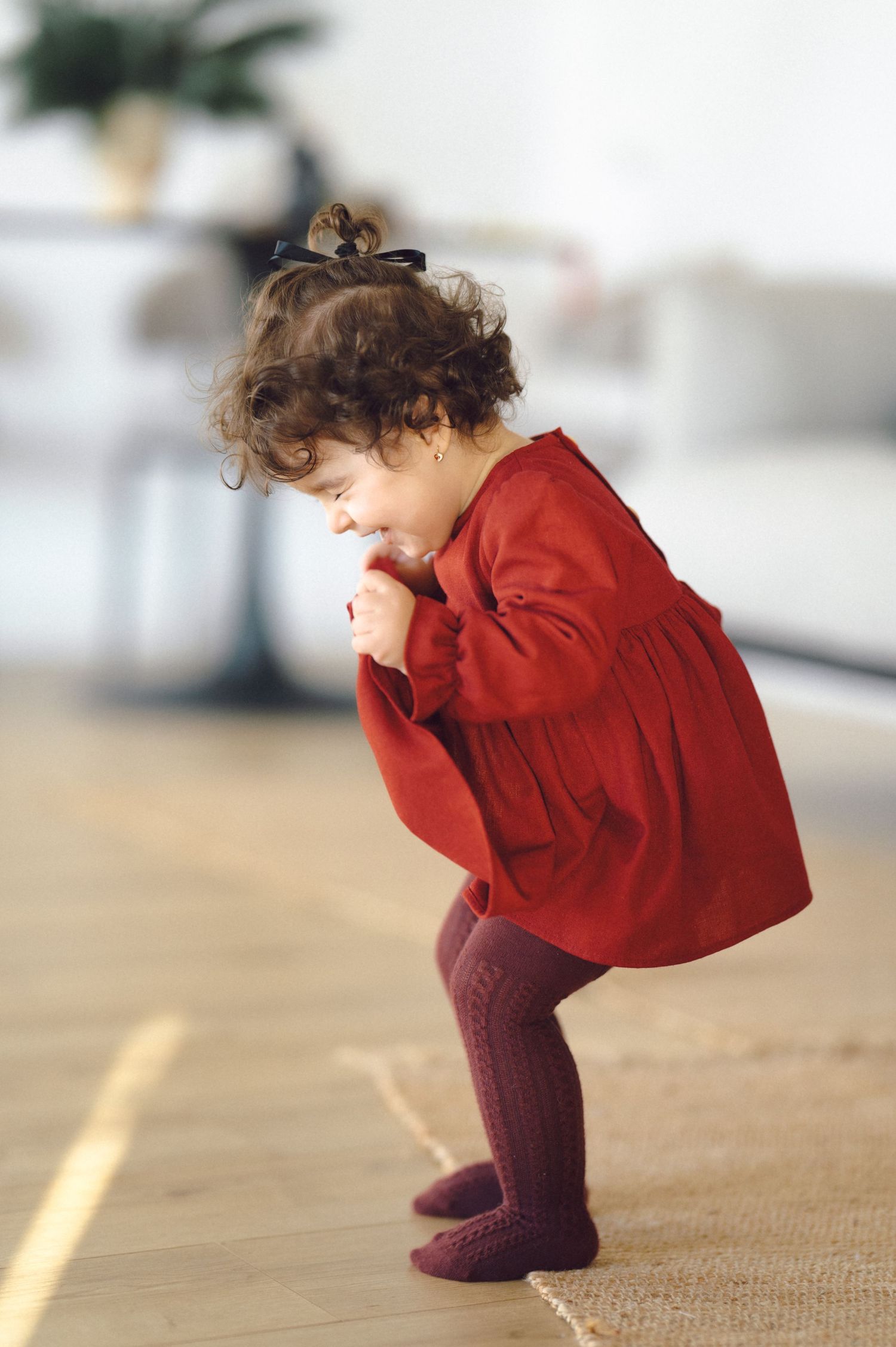A small child in a red sweater and burgundy tights leans forward curiously on a light wooden floor.