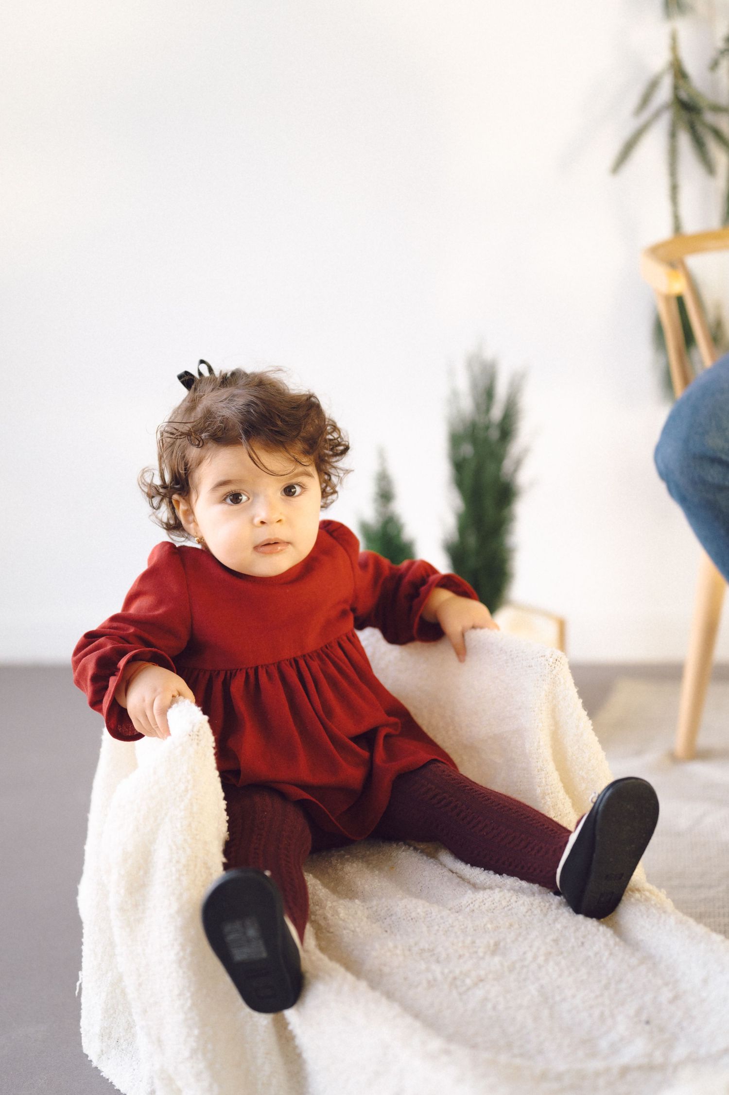 A toddler wearing a burgundy dress and tights sits on a fluffy white blanket against a minimalist winter backdrop.