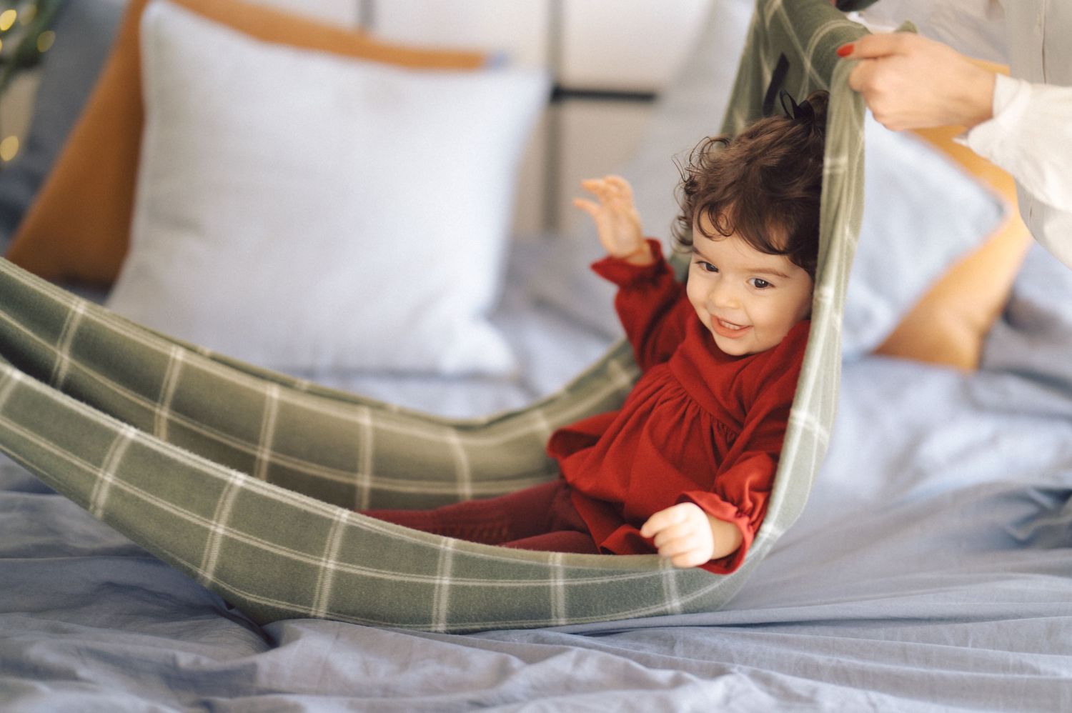A young child in a red sweater plays joyfully on a cozy bed with green plaid bedding.
