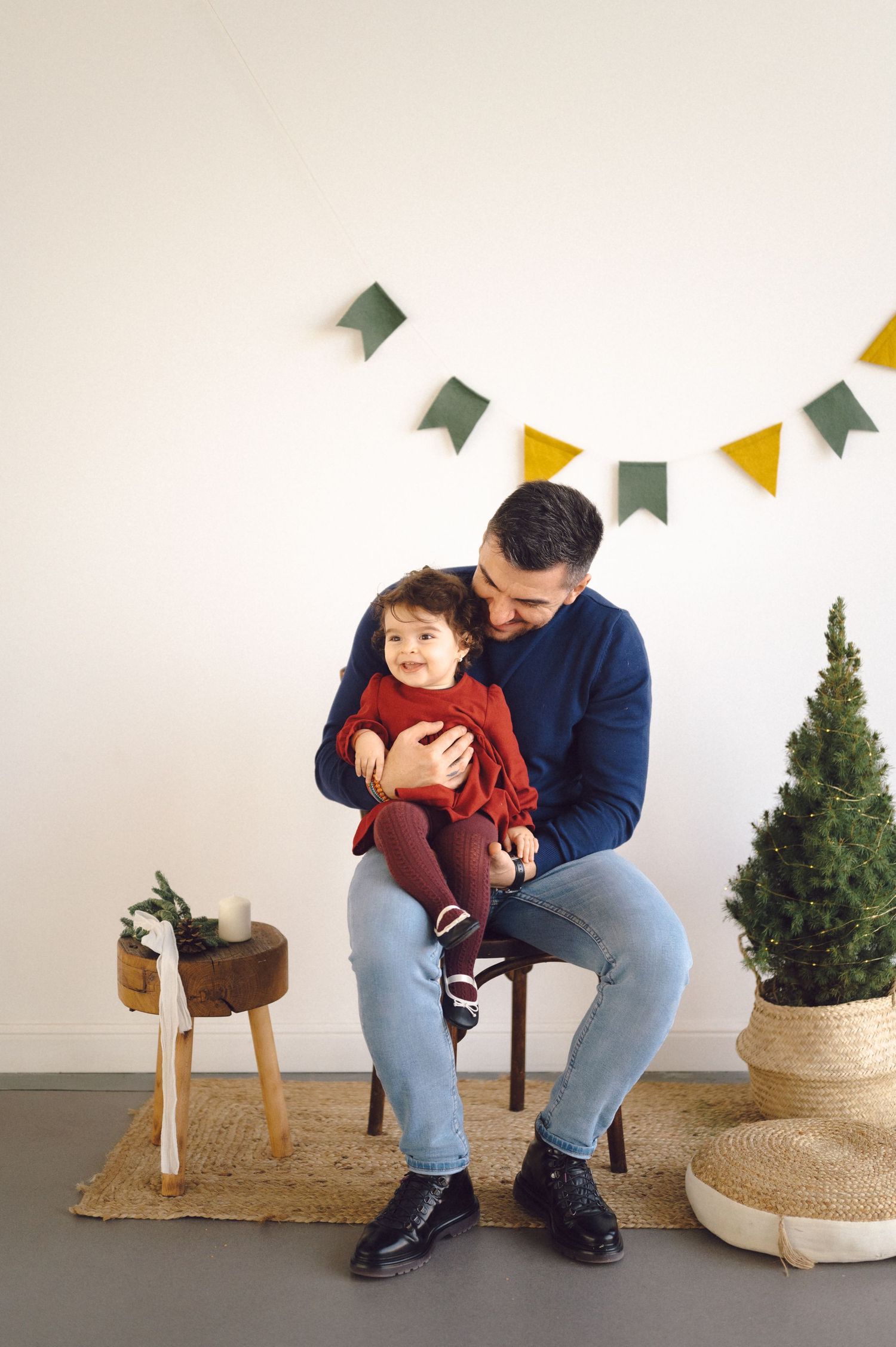 Festive indoor scene with bunting decorations, small potted trees, and a wooden stool beside people sitting in a chair.