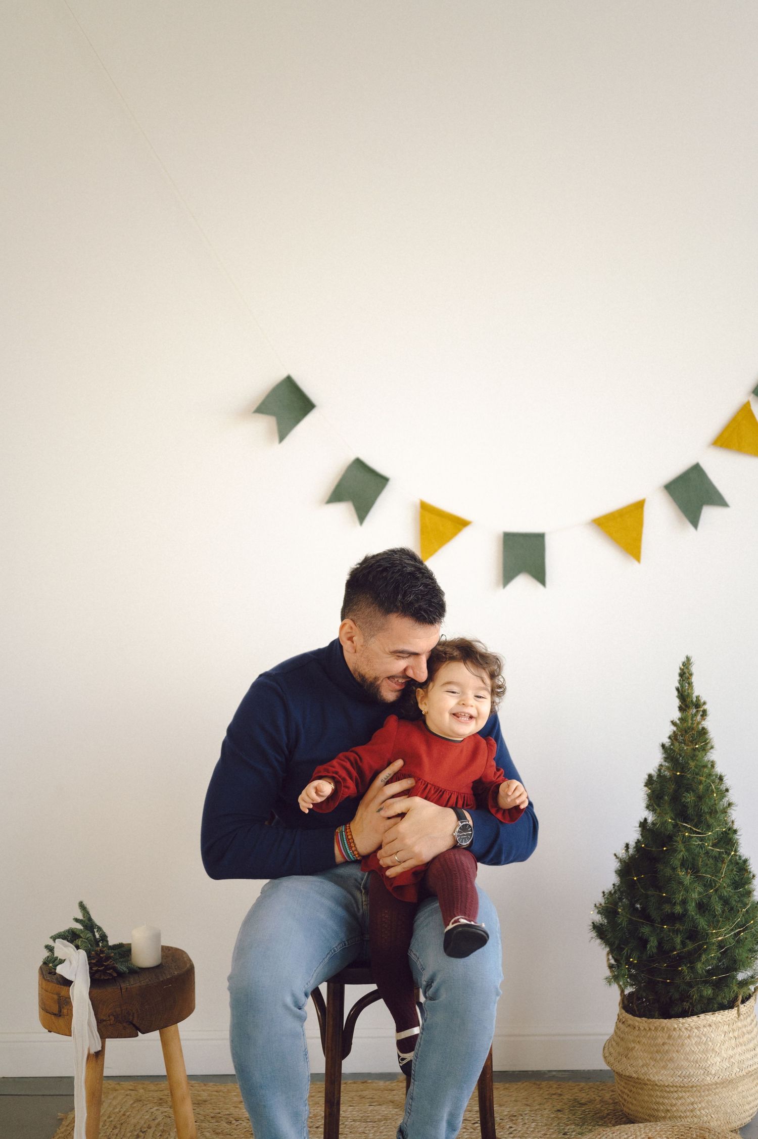 Cozy indoor scene with colorful bunting flags, a small potted tree, and a warm moment between parent and child on a stool.