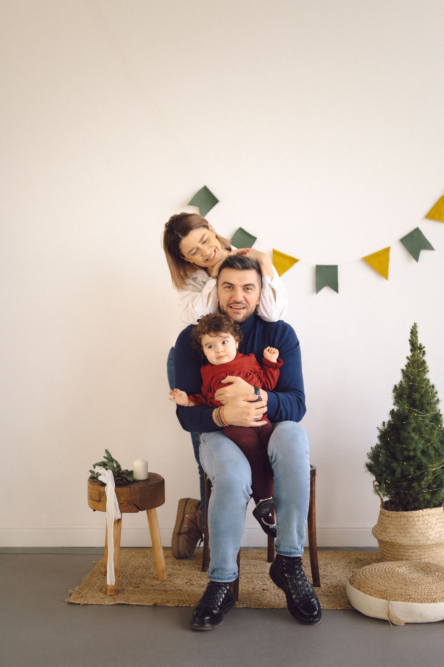 A parent and child sit on a small wooden stool next to a potted evergreen tree with yellow and green banners overhead.
