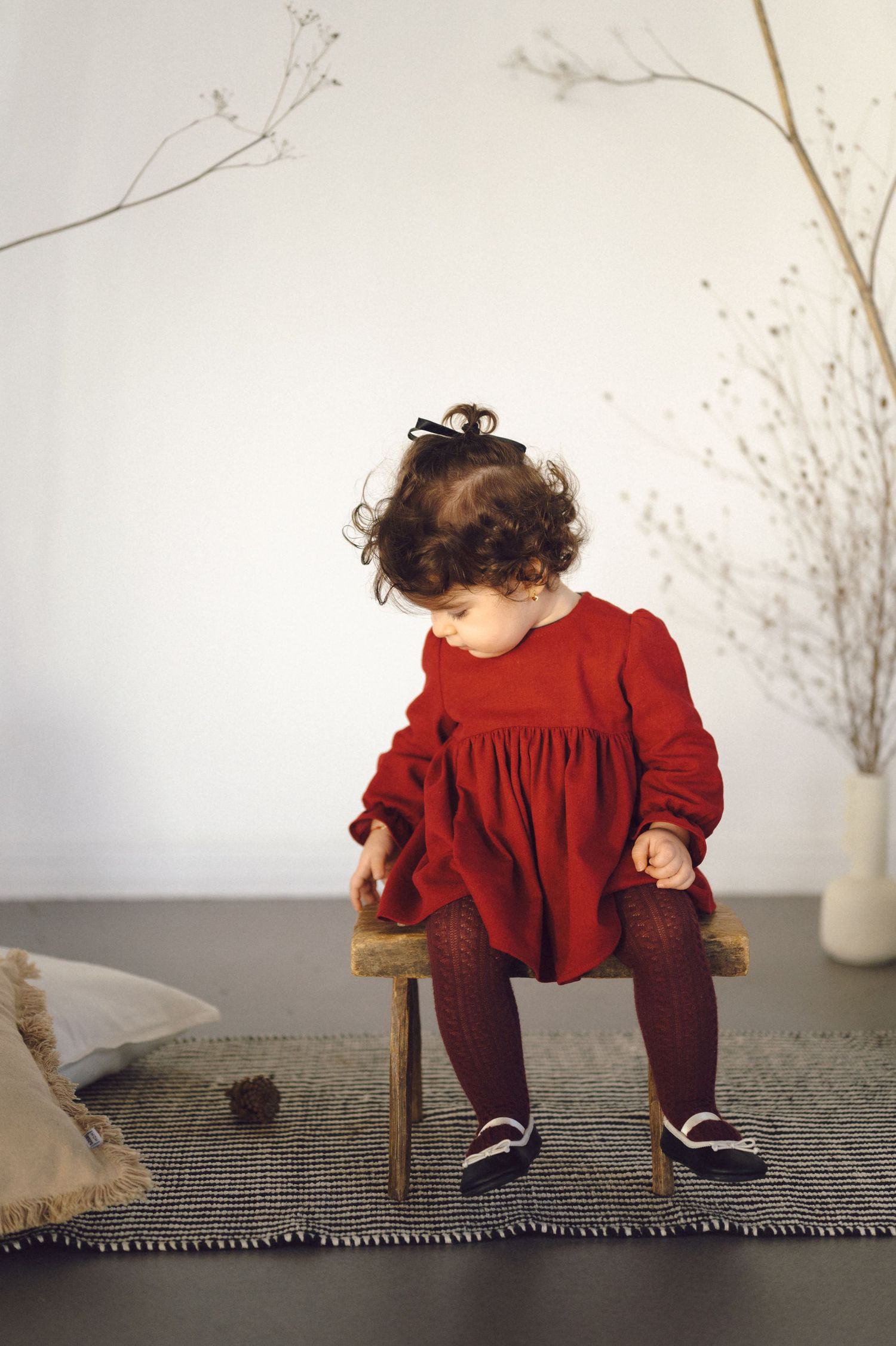 Child in red dress sits thoughtfully on textured surface against white background with delicate twigs in vase.