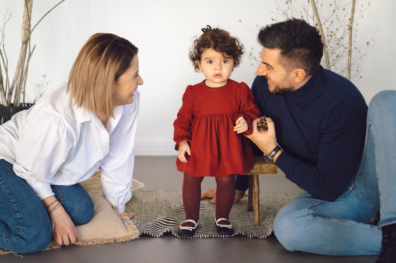 Two adults crouched down on the ground interacting with a small child wearing a red dress against a white wall.