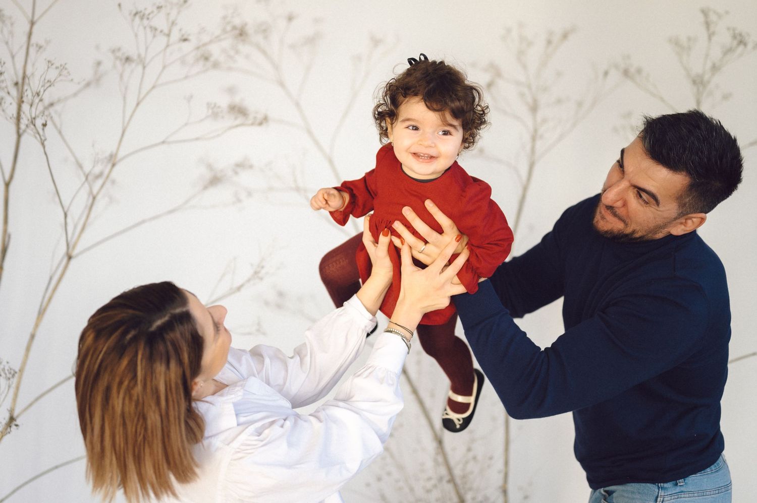 Parents in white and navy sweaters lift a smiling child wearing a red turtleneck sweater against a light background.
