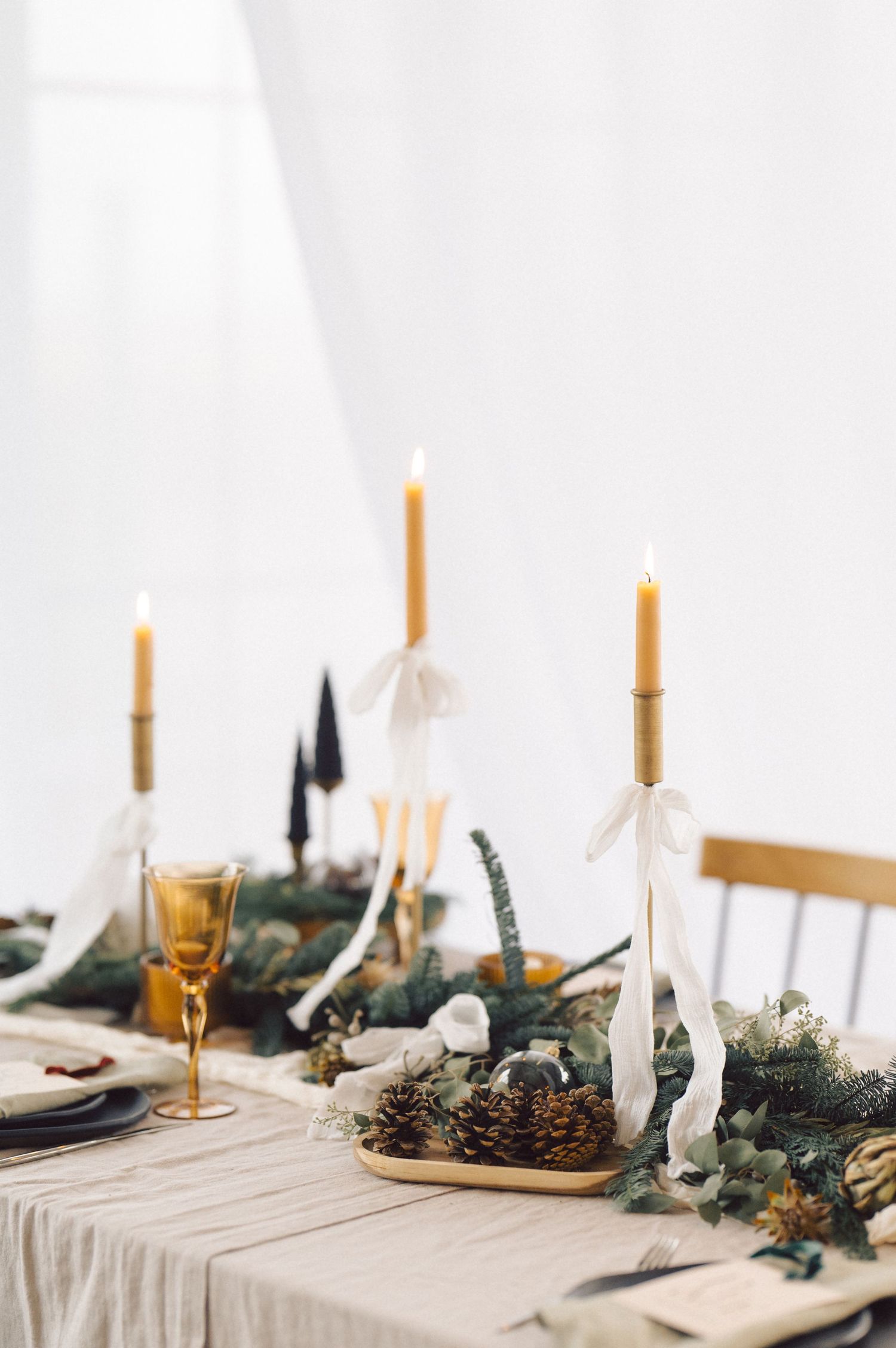 An elegant holiday table setting with tall gold candles, greenery garland, pinecones and champagne glasses on white linens.