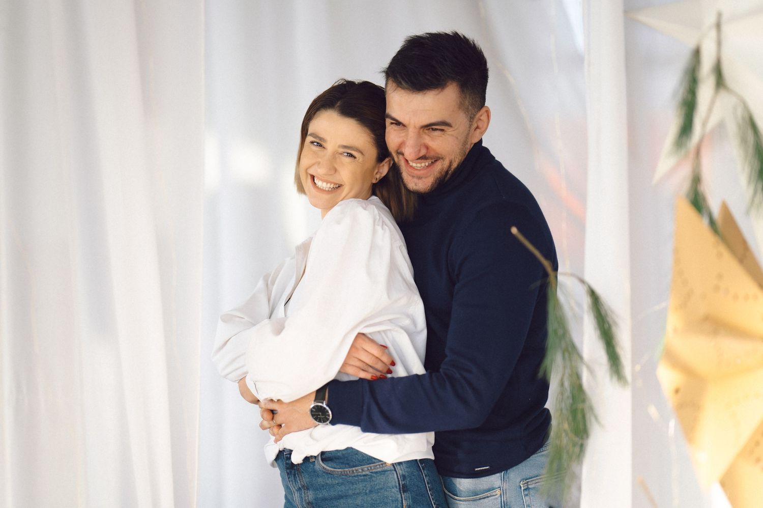 Two people in casual attire share a warm embrace while smiling and leaning against a white wall.