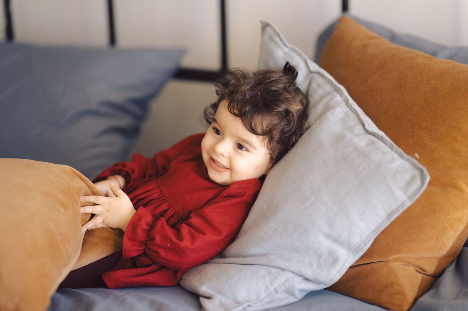 Small child lounges across a tan leather couch with grey pillows, wearing a burgundy long sleeve shirt.
