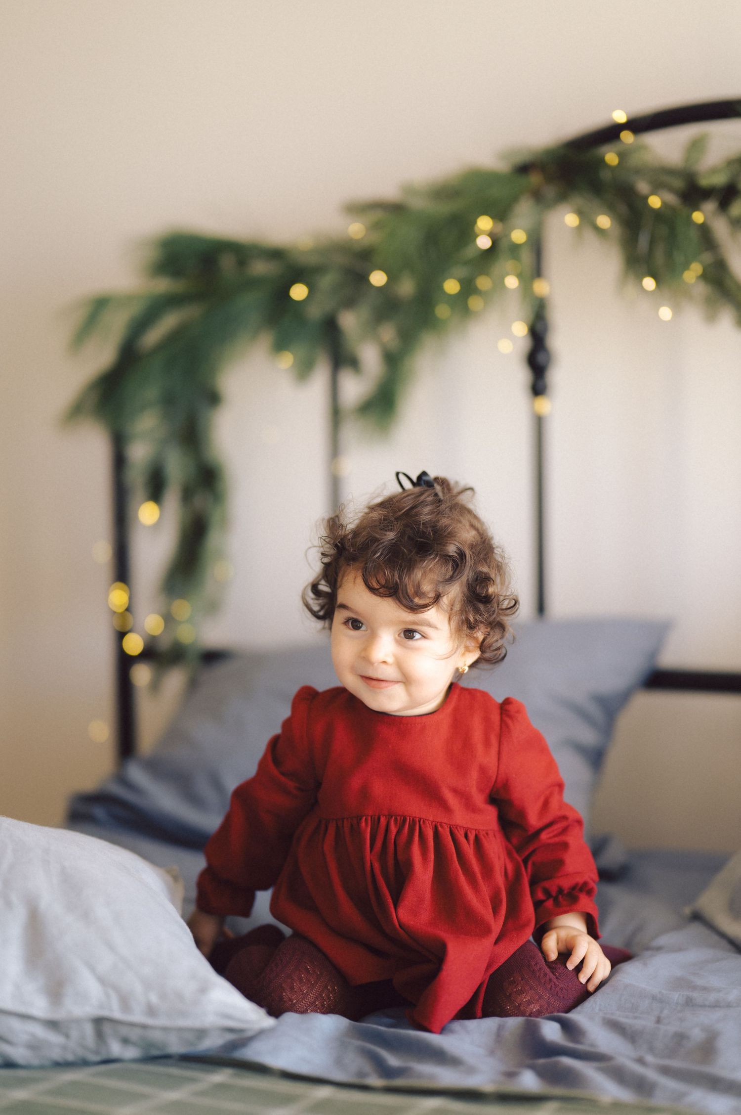 Small child wearing red dress with long sleeves sits on grey bedding with holiday decorations behind them.
