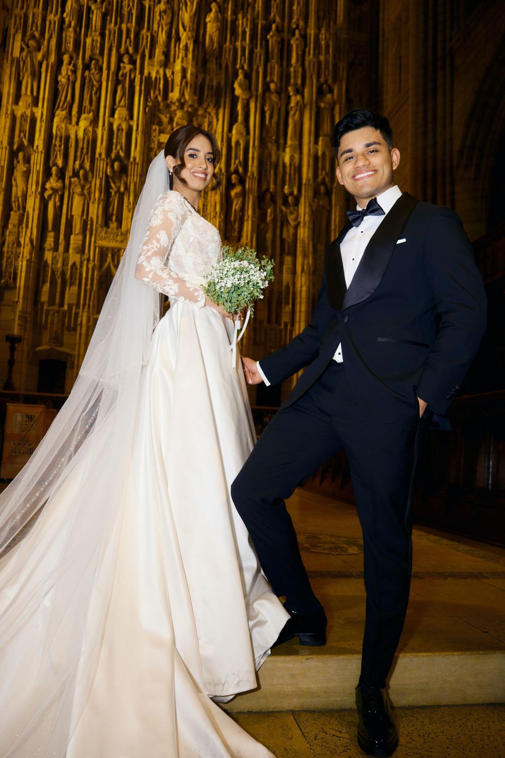 A bride in a flowing white gown and groom in black tux pose playfully in an ornate cathedral setting.