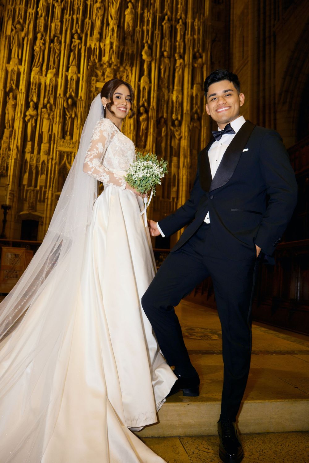 Newlyweds pose in a grand cathedral with ornate golden architecture, bride wearing a classic long-sleeved lace gown.