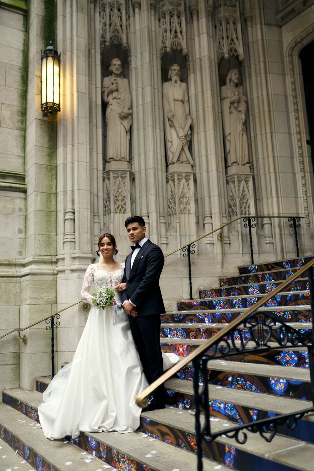 Wedding couple poses on decorated stone steps with Gothic cathedral architecture behind them.