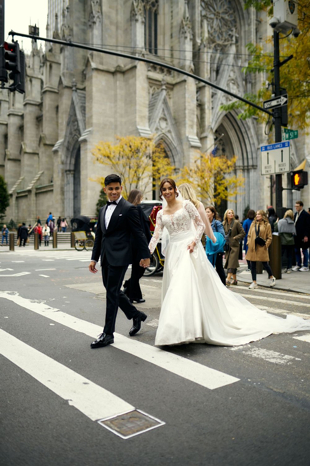 Newlyweds cross city street in front of majestic cathedral with fall foliage visible.